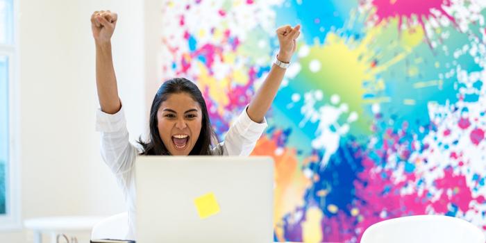 excited hr reading off a laptop screen with both arms in the air, multi-colored splashes of paint to the background